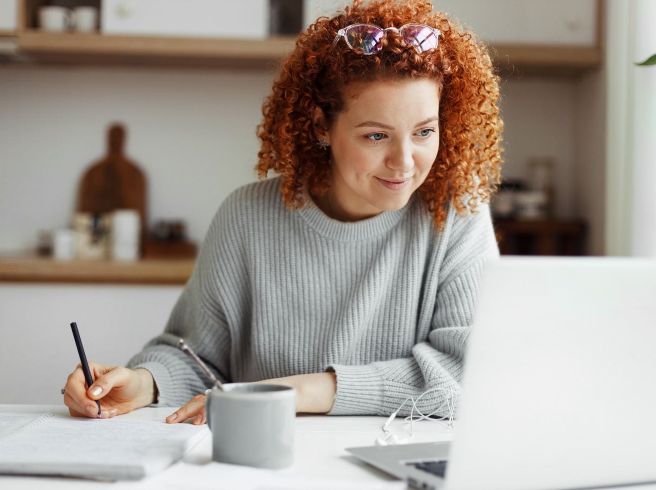 A woman with curly red hair writes in a notebook while looking at a laptop on a white desk, with a mug and earphones beside her. A woman with curly red hair writes in a notebook while looking at a laptop on a white desk, with a mug and earphones beside her.
