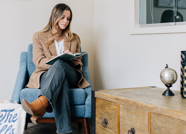 A woman sits on a blue chair reading a book, with a tote bag on the floor and a wooden cabinet with a small globe and decorative items nearby. A woman sits on a blue chair reading a book, with a tote bag on the floor and a wooden cabinet with a small globe and decorative items nearby.